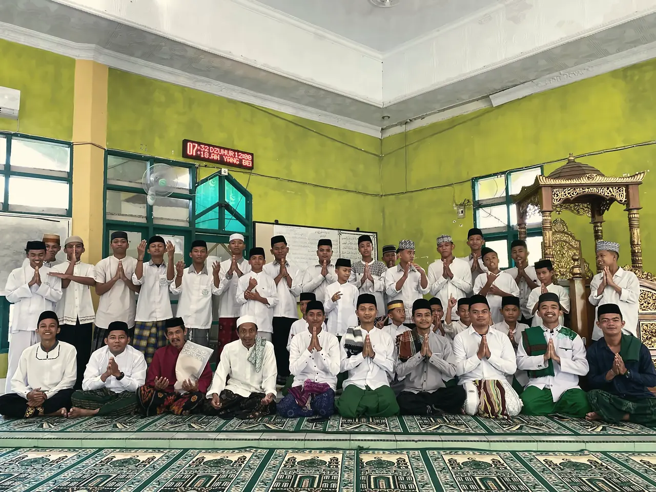 Para ustadz duduk di depan dan santri putra berdiri di belakang, berfoto bersama di masjid sambil menangkupkan tangan