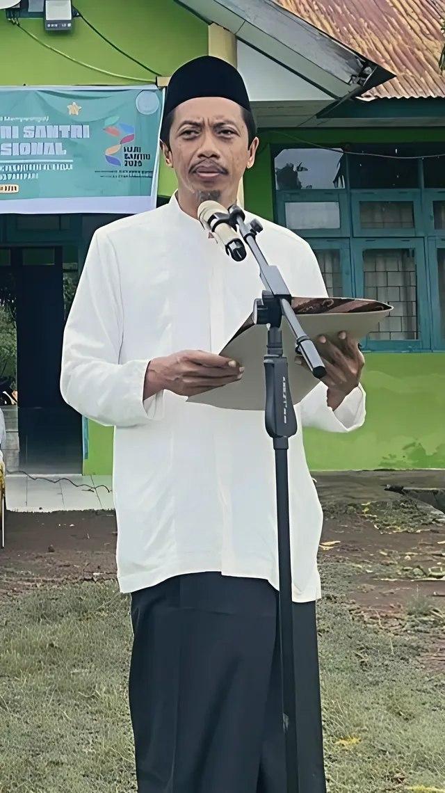 Seorang ustadz berpeci hitam membacakan teks di depan mikrofon pada upacara Hari Santri di halaman pesantren
