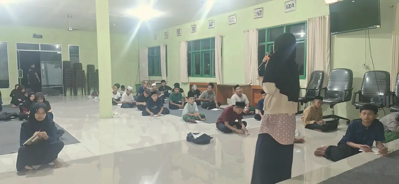 Santri duduk di lantai aula pada malam hari, mencatat pelajaran dari ustadzah yang berbicara dengan mikrofon