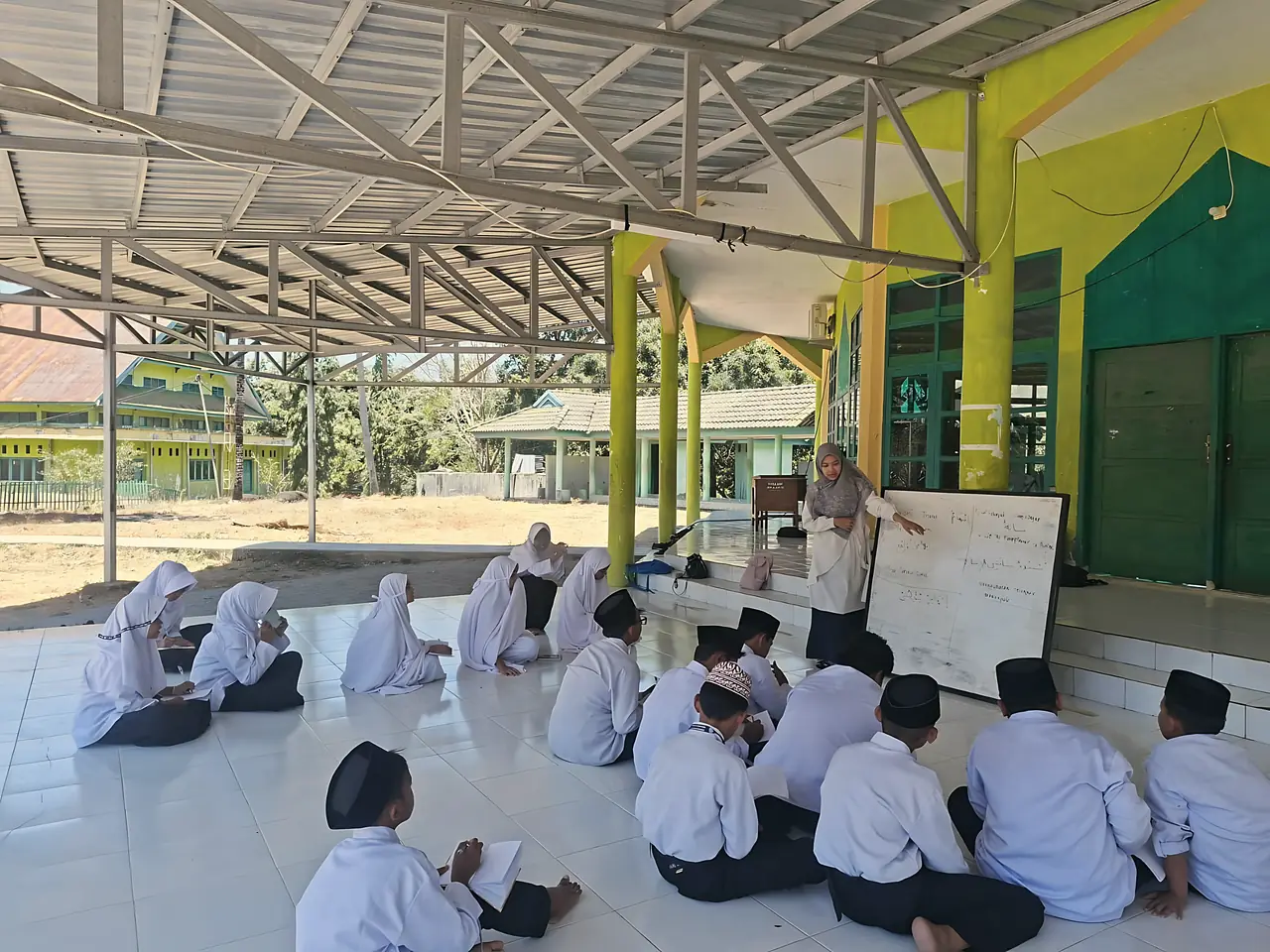 Ustadzah menjelaskan kosakata bahasa Arab di papan tulis kepada santri yang duduk di lantai serambi